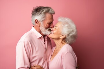 Senior couple, both in their late 70s, Caucasian, sharing a gentle kiss on the cheek against a soft pink pastel background with copy space
