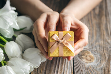 Small gift box in female hands on a wooden background with tulips.