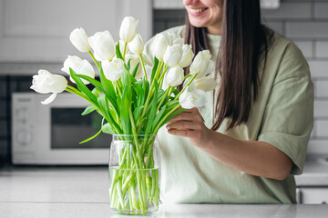 Happy Caucasian woman putting fresh white tulips into the vase. - obrazy, fototapety, plakaty