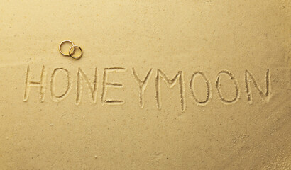Word Honeymoon written on sand and two golden rings, top view