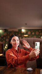 Smiling hipster girl waving hand hello during online video call via cellphone, sitting in cafe. Attractive business woman or student having conference in internet via smartphone. Copy space