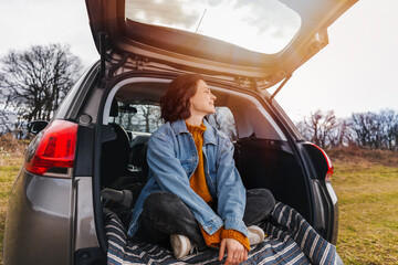 Young cheerful woman traveler sitting in open trunk of car enjoying nature& Solo travel concept