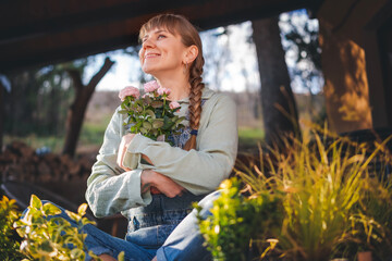 Happy young Caucasian woman with rose plant in a pot sitting on the terrace at cottage on a spring day. Flower seedlings for the garden and floriculture - 746382914