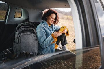 Young happy woman sitting in an open car trunk taking pictures with a camera. Traveling by car, communication in travel concept - 746383956