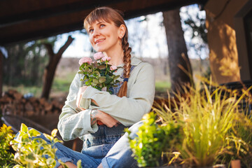 Happy young Caucasian woman with rose plant in a pot sitting on the terrace at cottage on a spring day. Flower seedlings for the garden and floriculture - 746384532
