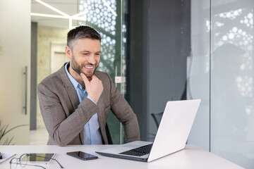 Smiling businessman in office using laptop with phone and tablet on desk