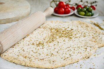 Focaccia dough with ingredients olives and cherry tomatoes on white table with rolling wood on the side.