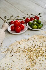 Focaccia dough with ingredients olives and cherry tomatoes on white table with rolling wood on the side.