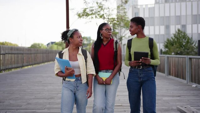 Happy African American nice women students walking carefree around university campus. Young black female friends stroll talking and smiling with backpacks and workbooks way to class spring day outdoor