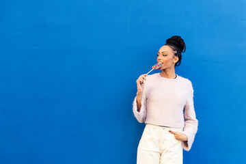 Smiling young woman eating lollipop in front of blue wall