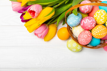 Easter basket filled with colorful eggs and a bouquet of tulips on a textured wooden table. Easter celebration concept. Colorful easter handmade decorated Easter eggs. Place for text. Copy space