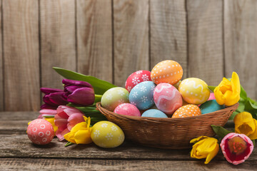 Easter basket filled with colorful eggs and a bouquet of tulips on a textured wooden table. Easter celebration concept. Colorful easter handmade decorated Easter eggs. Place for text. Copy space