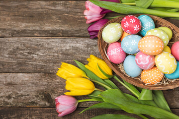 Easter basket filled with colorful eggs and a bouquet of tulips on a textured wooden table. Easter celebration concept. Colorful easter handmade decorated Easter eggs. Place for text. Copy space