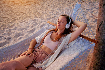 Young blissful calm woman wearing wireless headphones with closed eyes lie in a hammock and enjoy relaxing music