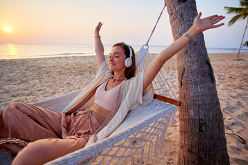 Young blissful calm woman wearing wireless headphones with closed eyes and raised arms lie in a hammock and enjoy relaxing music