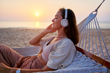 Young blissful calm woman wearing wireless headphones lie in a hammock and enjoy relaxing music