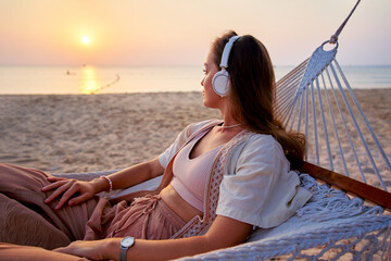 Young blissful calm woman wearing white wireless headphones lie in a hammock and enjoy relaxing music