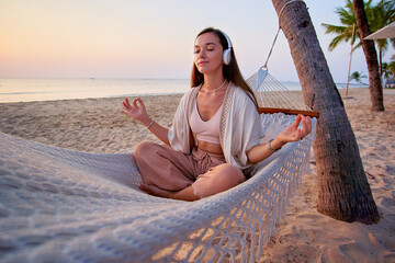 Young blissful calm woman wearing white wireless headphones with closed eyes and lotus pose hands meditates in a hammock and enjoy relaxing music, mind care