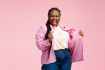Portrait overjoyed African American woman with stylish braids wearing trendy pink shirt isolated on background. Happy Nigerian plus size model dancing celebration success. Body positive concept