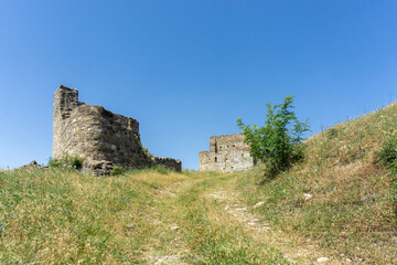 A dirt road leads to the ruins of medieval fortress on the hill. Stone wall and old building are visible. Blue bright sky.