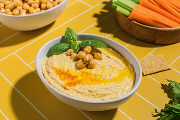 Delicious chickpea hummus in a plate on a bright yellow background with fresh vegetables. 
