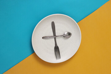 Empty plate with spoon and fork on a blue and yellow background, representing fasting during Ramadan and the anticipation of breaking fast