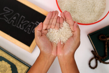 Hand holding a pile of rice for Zakat al-Fitr, with the Holy Quran, prayer beads or tasbih, and...