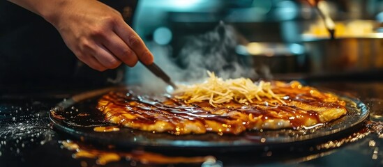 Hot traditional street food is prepared by a chef on an iron plate, where a Japanese pancake is topped with a salty sauce to enhance its deliciousness. - obrazy, fototapety, plakaty