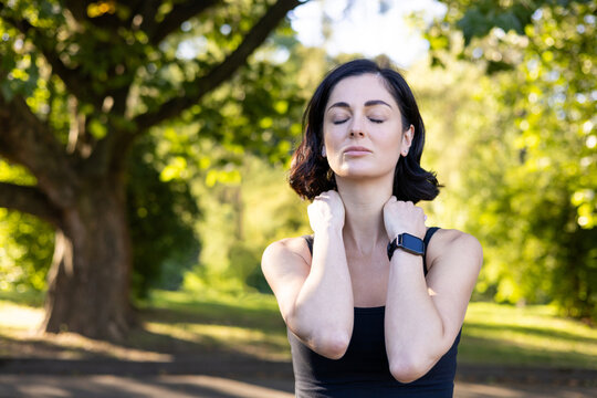 Close-up photo of young tired woman doing sports and exercising outdoors, standing exhausted with closed eyes, holding hands on neck, doing massage and resting
