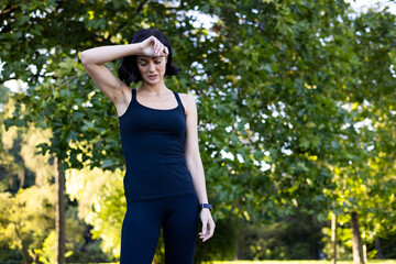 Young woman jogging and doing sports in the park, standing in sportswear and holding her head with her hand, feeling a headache and wiping sweat from her forehead when she is tired