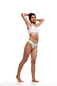Full-length image of beautiful young girl with tanned, slim, fit body posing in underwear against white studio background. Concept of body and health care, sport, female beauty, wellness