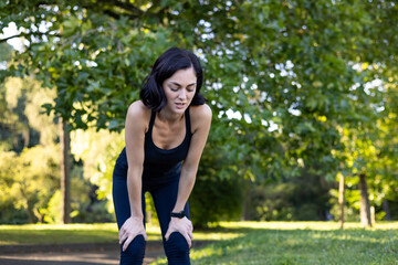 Tired young woman standing bent over in park, resting and shivering hard after jogging and training
