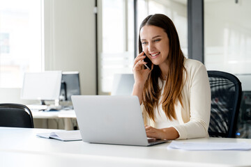 Young businesswoman is engaged in conversation on the phone while using laptop, illustrating the...