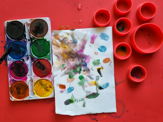 Toddler painting with watercolors. Workspace for kids' self-expression. Watercolor palette and a canvas with abstract painting. Kid's hand lays over his painting.