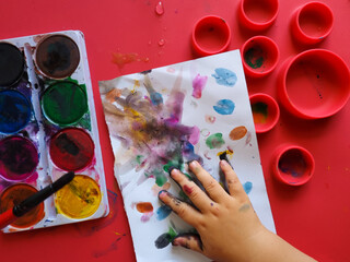 Toddler painting with watercolors. Workspace for kids' self-expression. Watercolor palette and a canvas with abstract painting. Kid's hand lays over his painting.