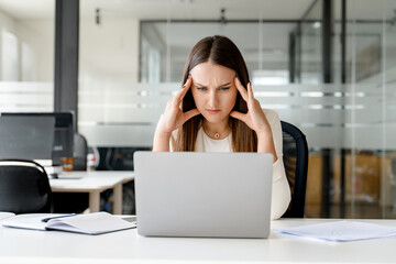 Stressed and concerned businesswoman holds head in hands, looking at a laptop screen, indicative of...