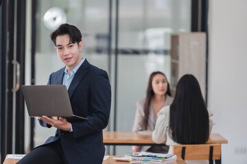 Smart businessman holding laptop while standing in the office room with his coworkers in the background.