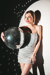 Fototapeta premium Young beautiful smiling blond female in trendy evening silver dress. Sexy carefree woman posing near white wall in studio in a circle of light. Fashionable model with bright makeup. Holds disco ball