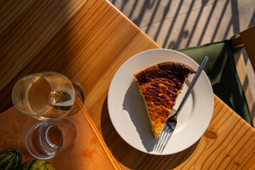 a slice of quiche on a white plate with a fork beside it, accompanied by a glass of white wine. The...