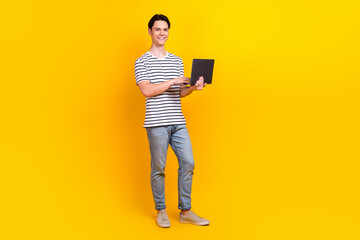 Full size photo of smart student dressed striped t-shirt denim pants hold laptop browsing eshop isolated on yellow color background