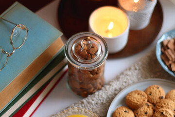 Cup of tea or coffee, stack of books, e-reader, glasses, various cookies, almonds, orange juice and lit candles on the table. Hygge at home. Selective focus.