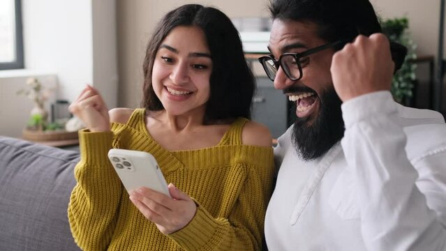 Surprised Indian couple receiving good news, exclaiming wow and giving high five, celebrating success or online victory with phone, relaxing on sofa at home living room.