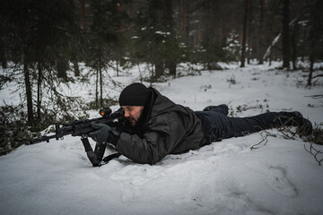Fototapeta na wymiar A private military man, in a prone position, aims from an AK-12 assault rifle in the forest in winter, side view.