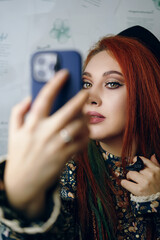 Red-haired girl takes a selfie on the front camera of a modern smartphone. Female blogger shoots herself for a blog about fashion and makeup. Adorable woman with a ethnic slavic crown on her head.