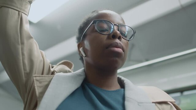 Low angle close up of African American 30 yo lady in glasses fixing earbuds while travelling by public transport