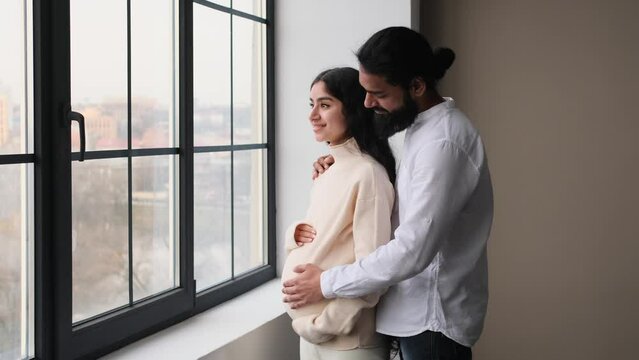 Indian couple, wife and husband, stands by the window at home, embracing and strokes pregnant belly, anticipate future parenthood, enjoying new baby expecting.