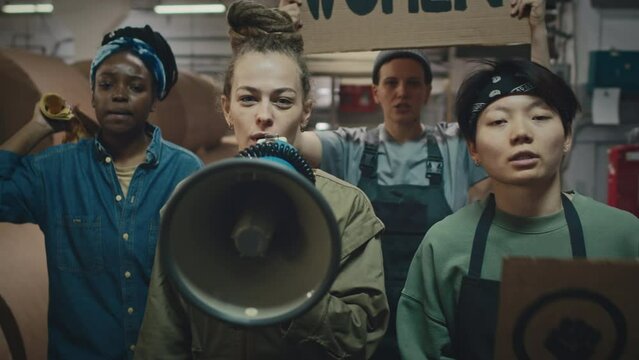 Team of young supportive multiethnic women with mouthpiece and feminist cardboard banners walking along industrial factory and showing their protest determinately