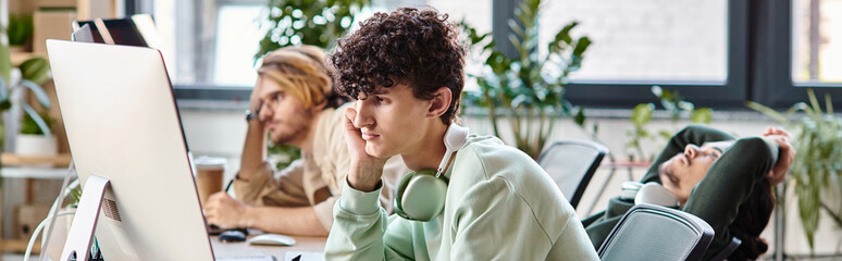 Young curly-haired man editing photos on tablet and looking at monitor in startup office, banner