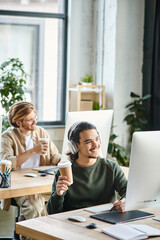 cheerful male professional in headphones holding coffee to go and using graphic tablet in office