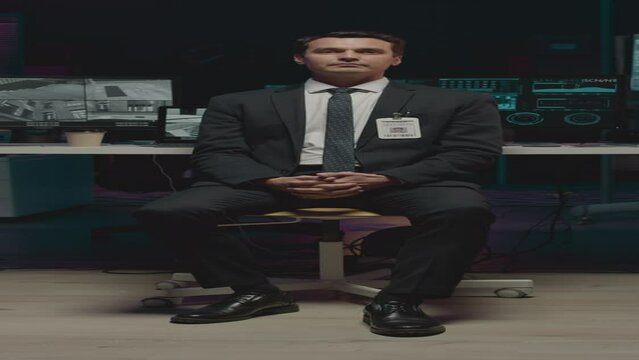 Vertical full portrait of Caucasian male cybersecurity officer in formalwear sitting in office chair by desk with CCTV cameras on computer monitor, adjusting his badge and looking at camera